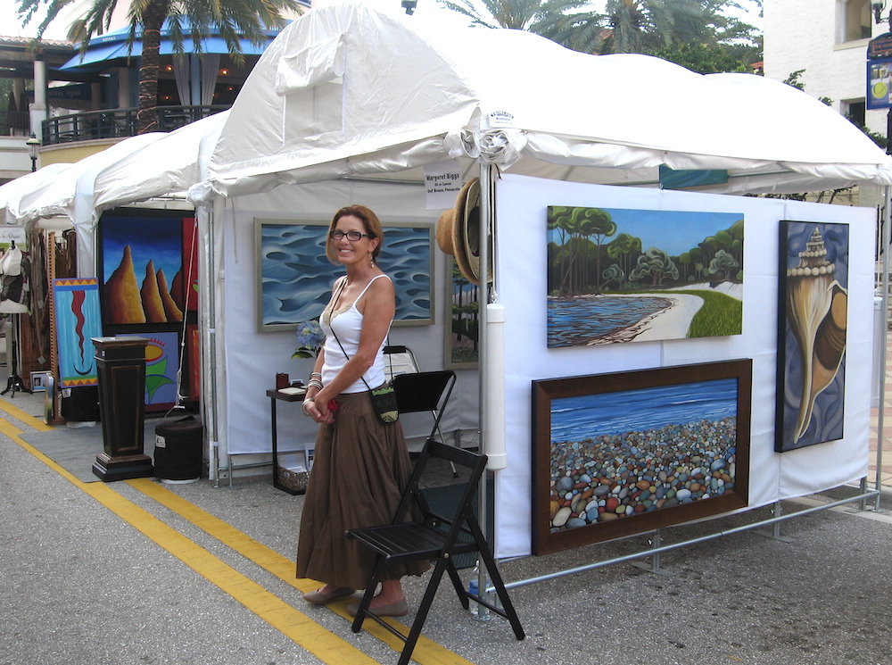 Margaret Biggsstands in front of a white tent full of hanging paintings | on Art Biz Success