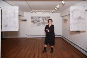 Lori Sokoluk in a black dress stands in the middle of a gallery with black and white work on wall | on Art Biz Success
