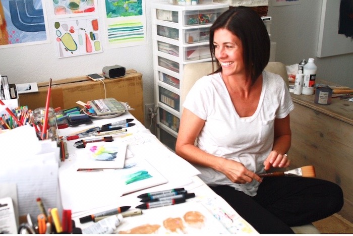 Sara Schroeder artist sitting in her studio chair at a work table filled with art supplies | on Art Biz Success