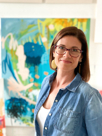 Photo of Sara Schroeder with glasses and a denim shirt and white t-shirt standing in front of her colorful abstract painting | on Art Biz Success