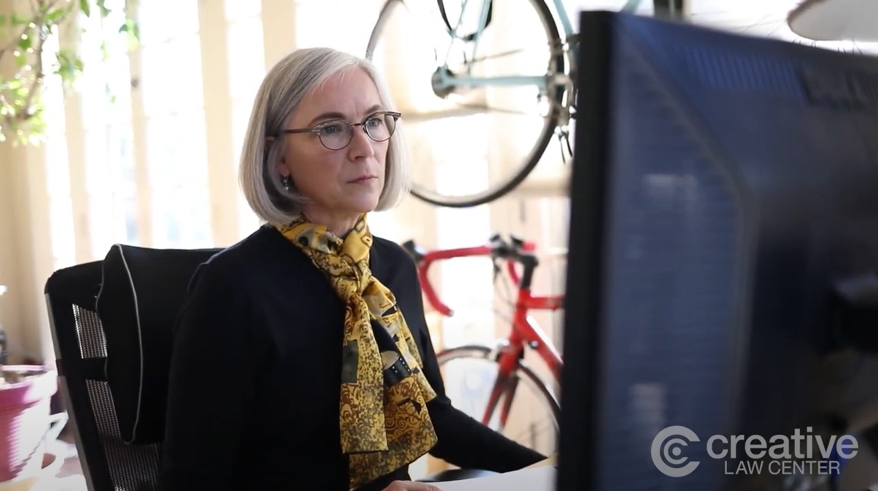 woman with gray hair wearing black jacket and yellow scarf at computer Kathryn Goldman | on Art Biz Success