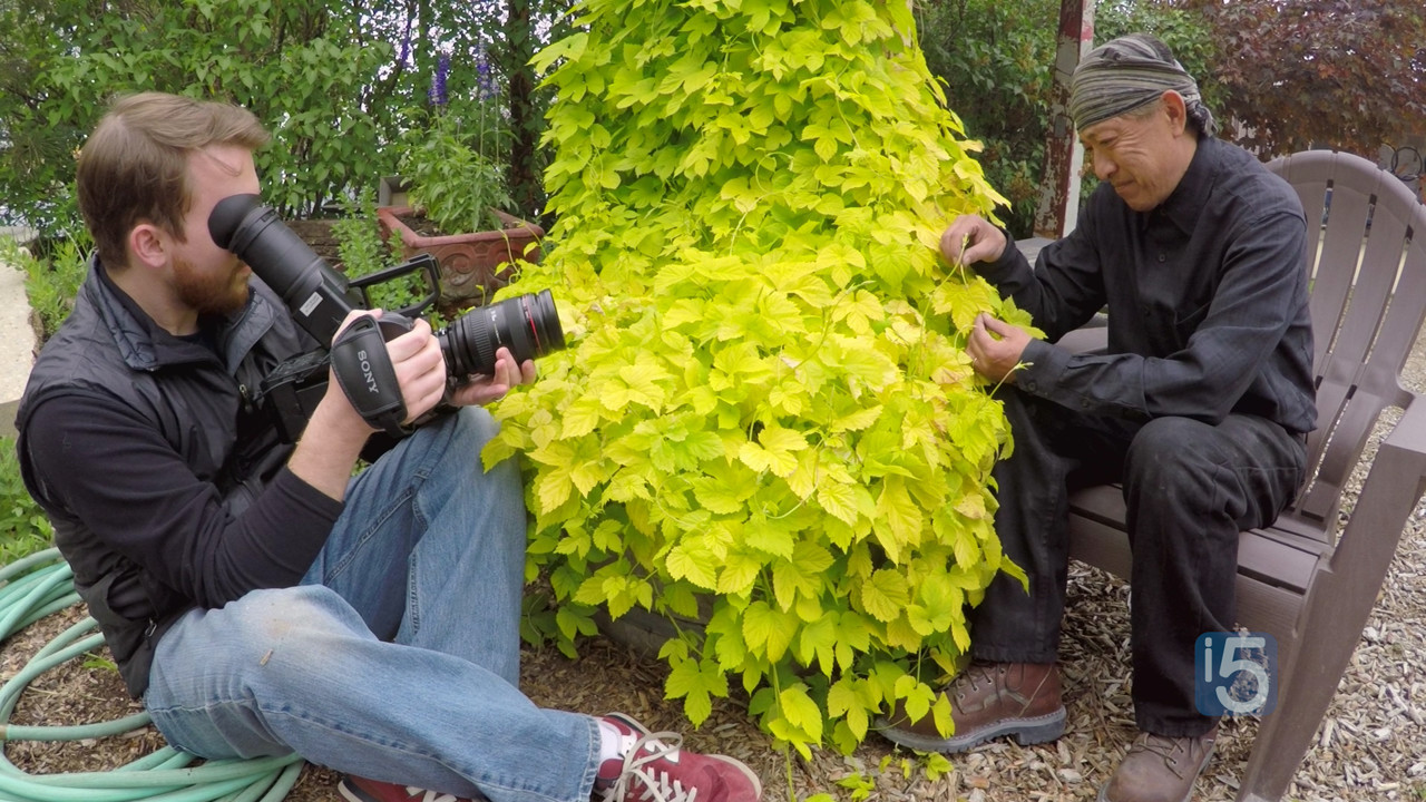 seated white man holding video camera pointed at man seated pruning green bush