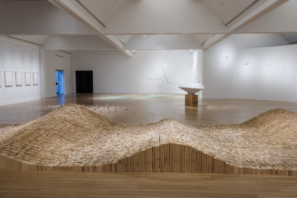 View of a rolling wood sculpture installation on the floor inside a white gallery space