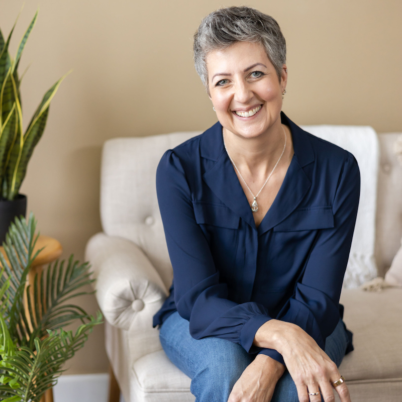Georgie Coote in a navy top sitting on a white couch with her hands crossed on her knees. Plants are on the left side.