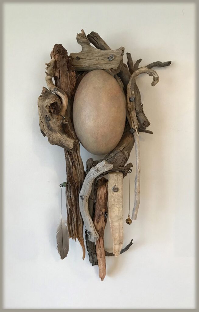 Monochromatic wall sculpture made of found wood, a feather, and held together with spiral-shaped metal pieces. A ceramic egg shape is nestled in the center.