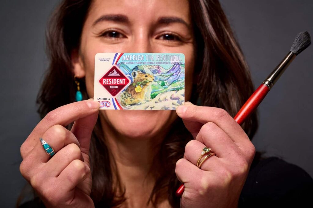Woman with dark hair holding up a National Parks pass with a painted scene adhered to the top and a paintbrush in her hand.